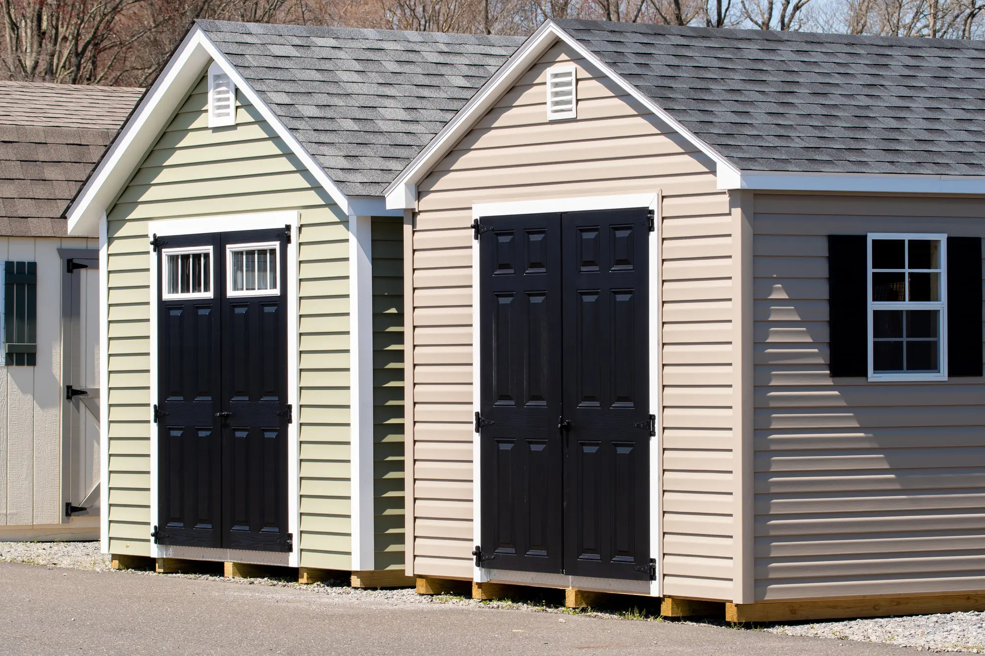 Two tan storage sheds with black doors and shutters side by side on a gravel lot