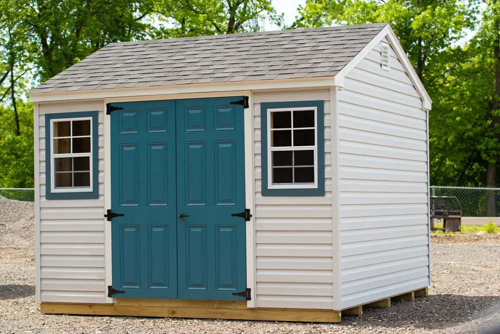 White storage shed with a blue door, blue shutters and double windows on a gravel pad